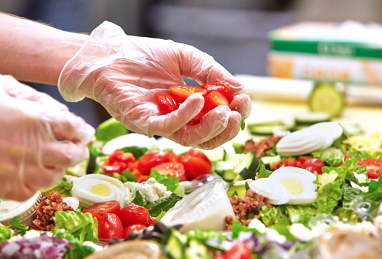 A caterer wearing plastic gloves picks up chopped tomatoes from a platter of fresh salad.